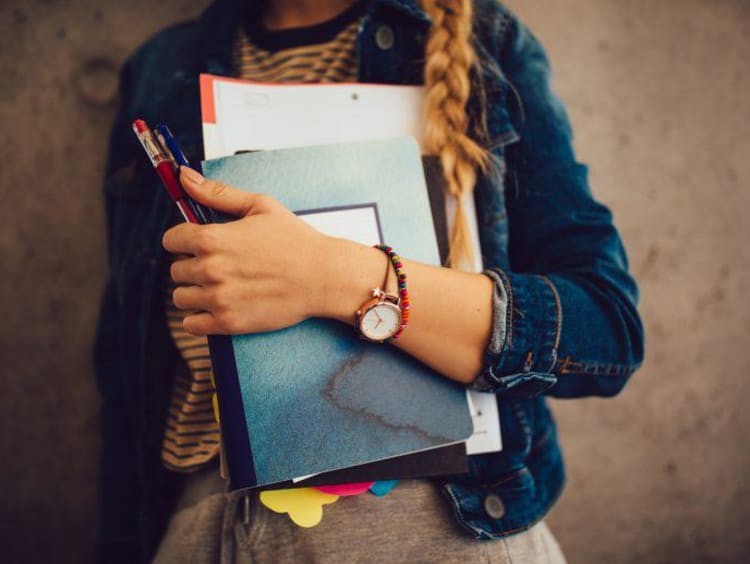 Teenage girl holding books, notebooks and pencils standing against wall