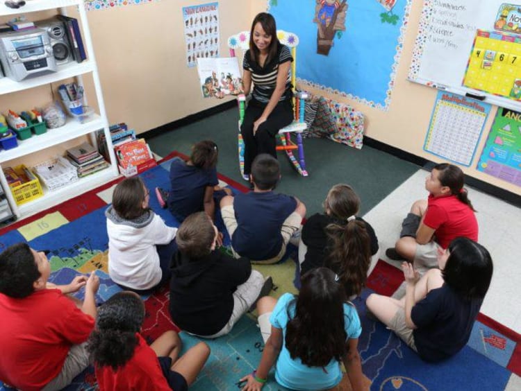 teacher sitting on chair reading book to students sitting on floor around her