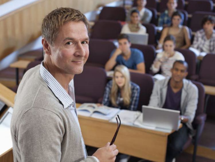 Male instructor stands next to podium with a full auditorium of students