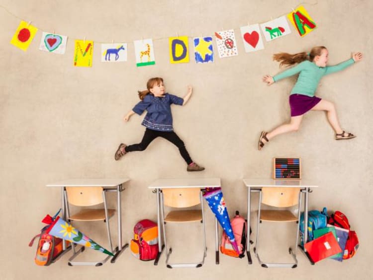 Two girls jump over desks next to a wall