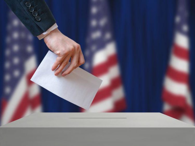 Ballot being placed in voter box with American flags behind the voting station.