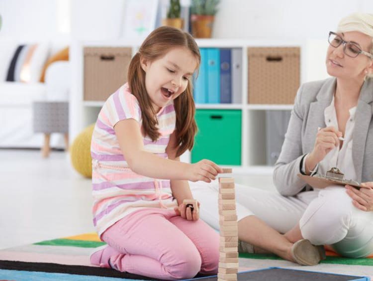 Child counselor sits on floor and observes a little girl playing with wood blocks