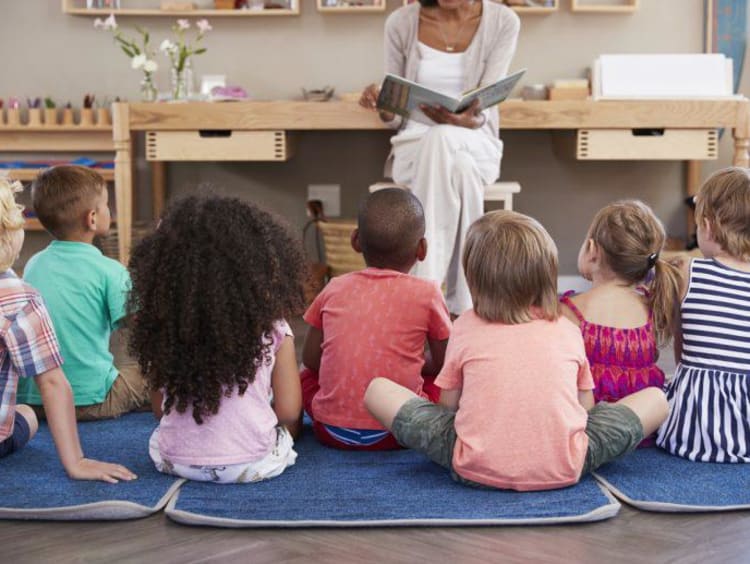 Elementary teacher sits at front of class reading a book to children sitting on ground