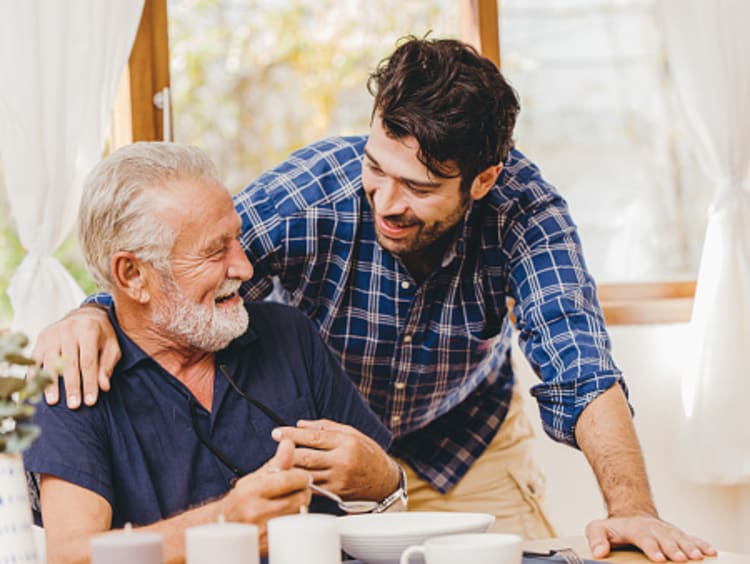 Man in plaid shirt demonstrates caring for others by putting his hand on an older gentleman seated at a table