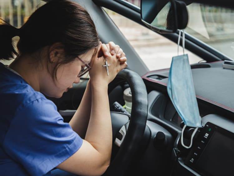 Female brunette nurse in the car praying with mask hanging from rearview mirror