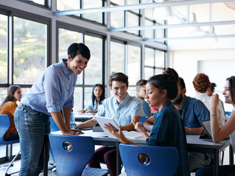 Smiling public school teacher leans on table to listen to student ask question when reading her tablet