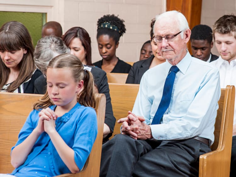 Community church pews full of people of all ages and races with heads bowed and hands clasped