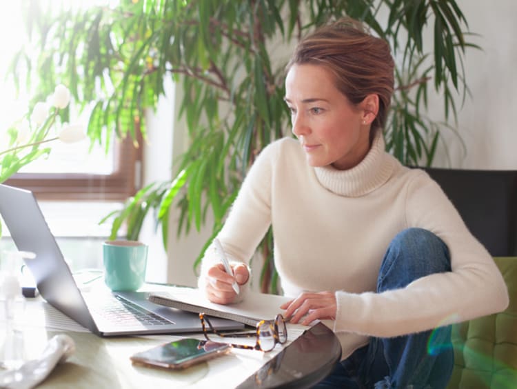 Slender blonde woman takes notes to develop her creative writing story on her laptop