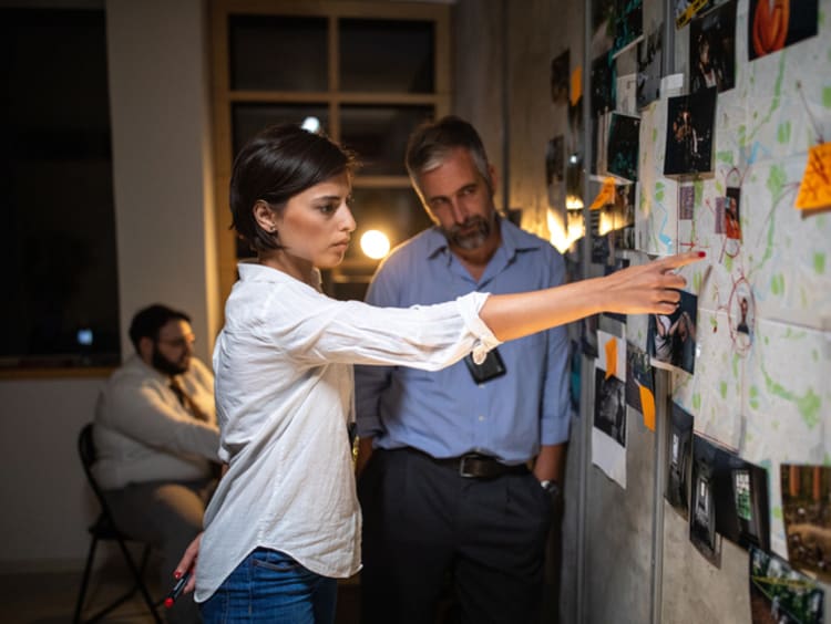 Woman police detective showing location on map attached to poster on wall to colleague