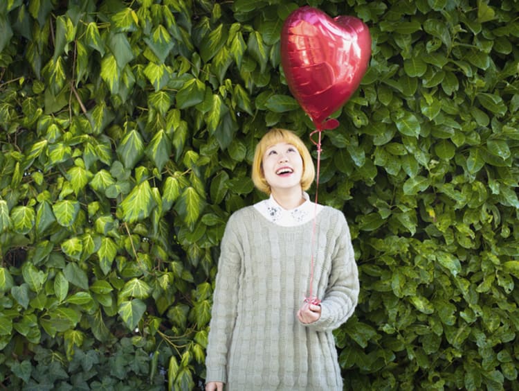 Asian woman with a blonde wig looks up at the heart-shaped balloon she's holding