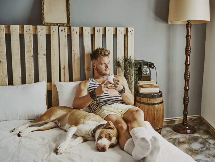 Man holds coffee cup looking thoughtfully out the window with dog snuggled up to him on the bed