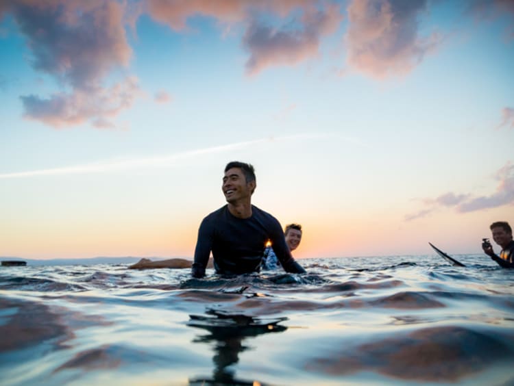 Man smiling while taking in the sunset in the ocean with other surfers