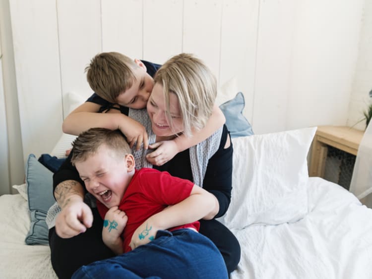 A young son kisses mom on the cheek on her bed and his brother is laughing