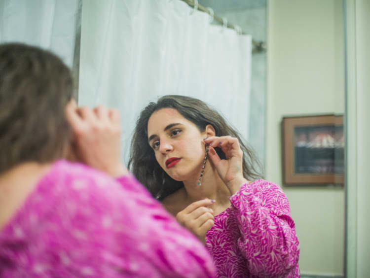 Caucasian brunette woman in pink dress putting earring on while looking in a mirror