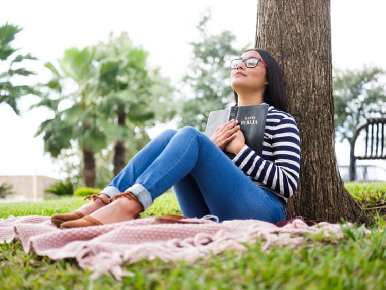 Woman holding an open Bible close to her chest and praying under tree