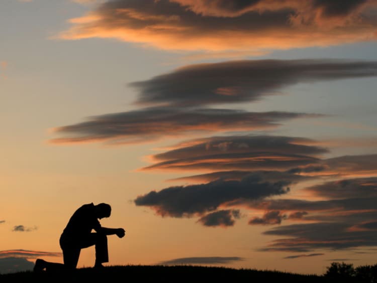 Illumination of a man kneeled on top of a hill praying in front of an ending sunset