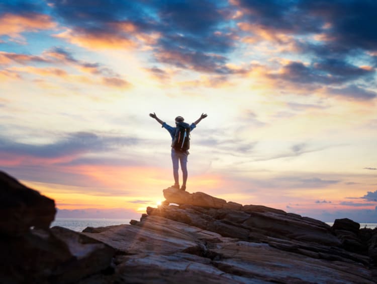 Person stands triumphantly on top of a mountain with sunset in horizon