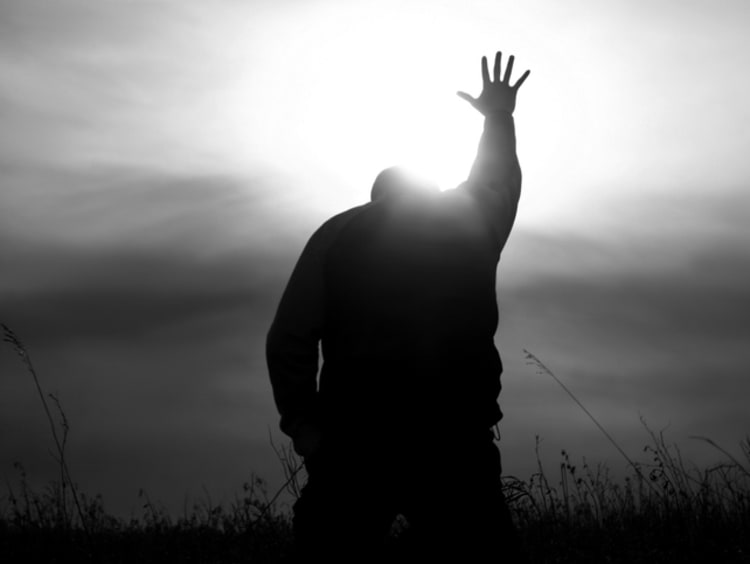 Black and white image of a man on his knees with one arm and hand raised up towards the sun