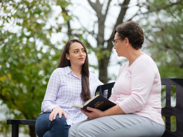 Older woman with Bible open in lap encourages younger woman listening to her