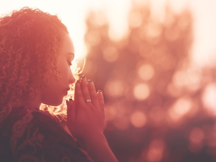 Woman praying outdoors with hands clasped as the sun sets