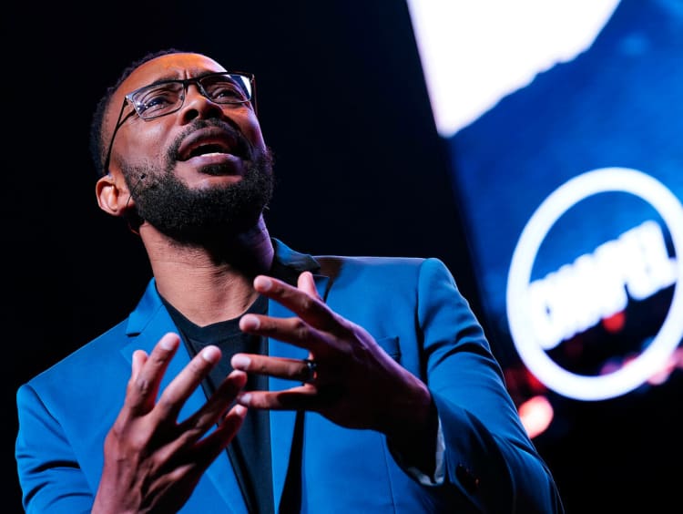 Marcus Doe speaking at GCU chapel - African male in a blue suit with black frame glasses expresses a point at a chapel service