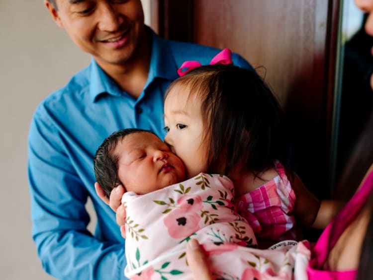 Little Asian girl kissing baby sister on the cheek while man in blue shirt watches