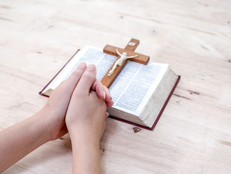 Praying hands rest at the foot of an open Bible