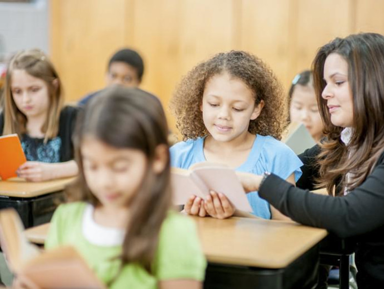 Kids in reading class and teacher kneels to assist a student with a word