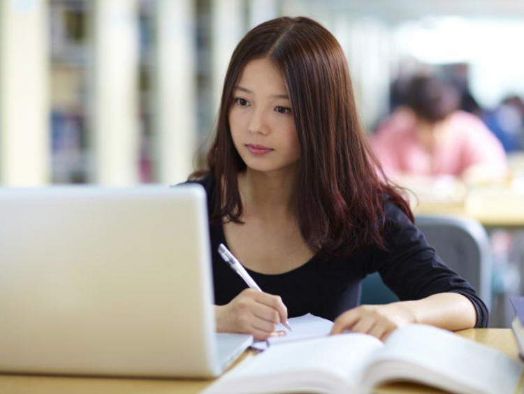 An Asian psychology student handwrites notes from laptop screen