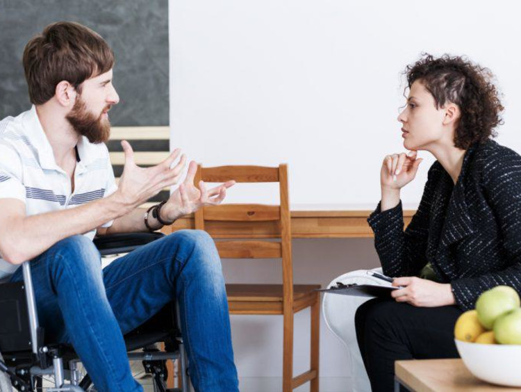 Man in wheel chair is talking and using his hands while a female trauma counselor leans in to listen