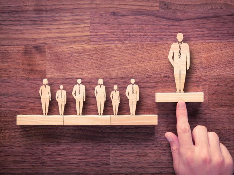 Wood outlines of people in a row with one being pushed higher by a real hand