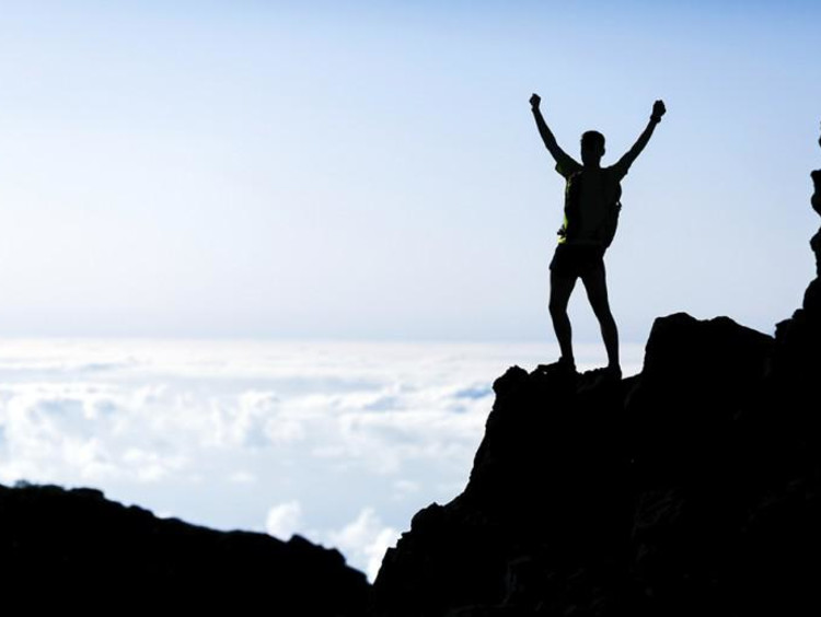 Man stands on top of mountain cliff raising hands in triumph