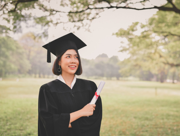 graduate standing under a tree in their cap and gown holding their degree