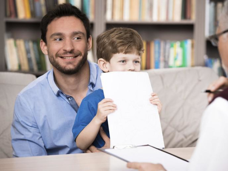 Father holds son who covers his face with papers while male counselor talks