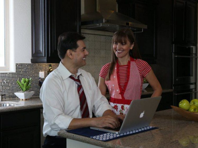Husband uses laptop in kitchen and wife in apron talks to him