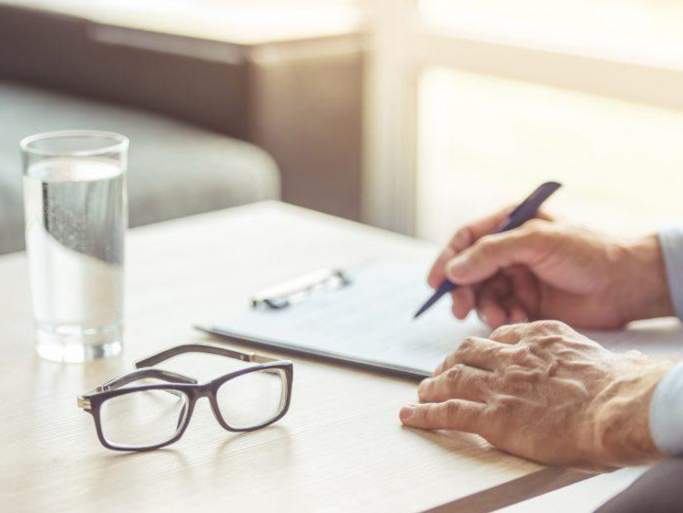 Glass of water and pair of eye glasses on desk while an older male uses checklist