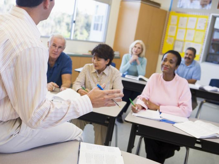 Male teacher sits on desk facing his class of adult students
