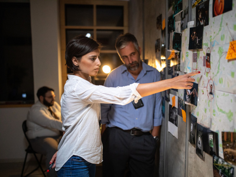 Woman police detective showing location on map attached to poster on wall to colleague