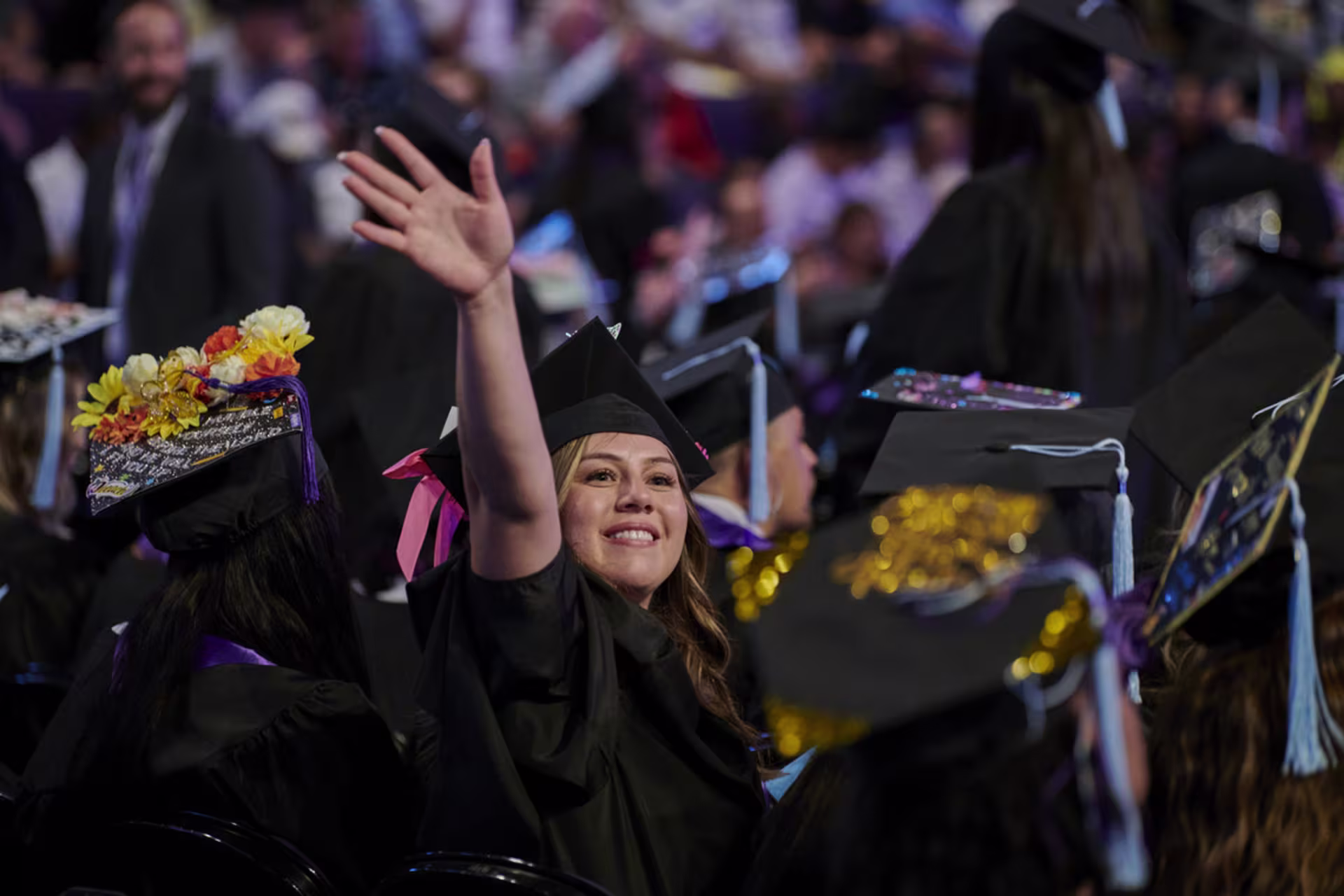 excited female graduate waves to her family amongst sea of seated graduates