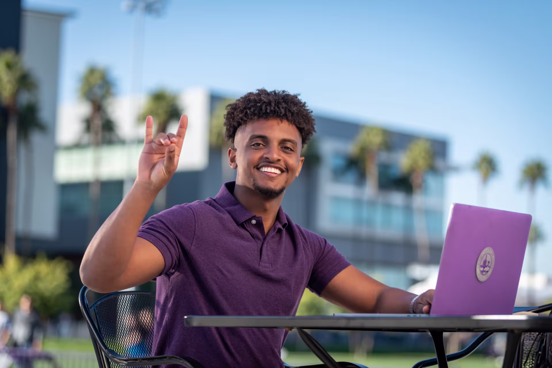 Student sitting outdoors on GCU campus with a laptop, raising a hand in celebration, representing starting a public health degree program at GCU.