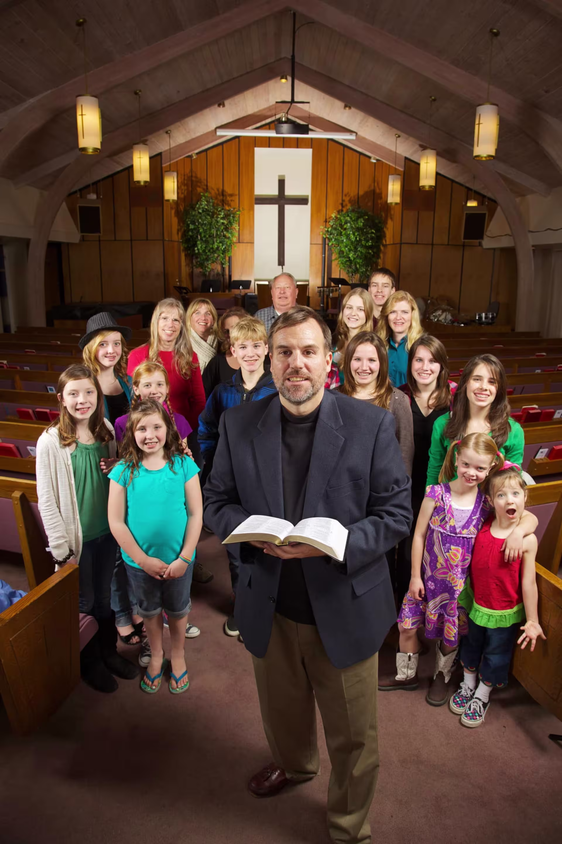Group of children, teens, and adults, including a pastor with a Bible, in a church setting, highlighting community in youth ministry.