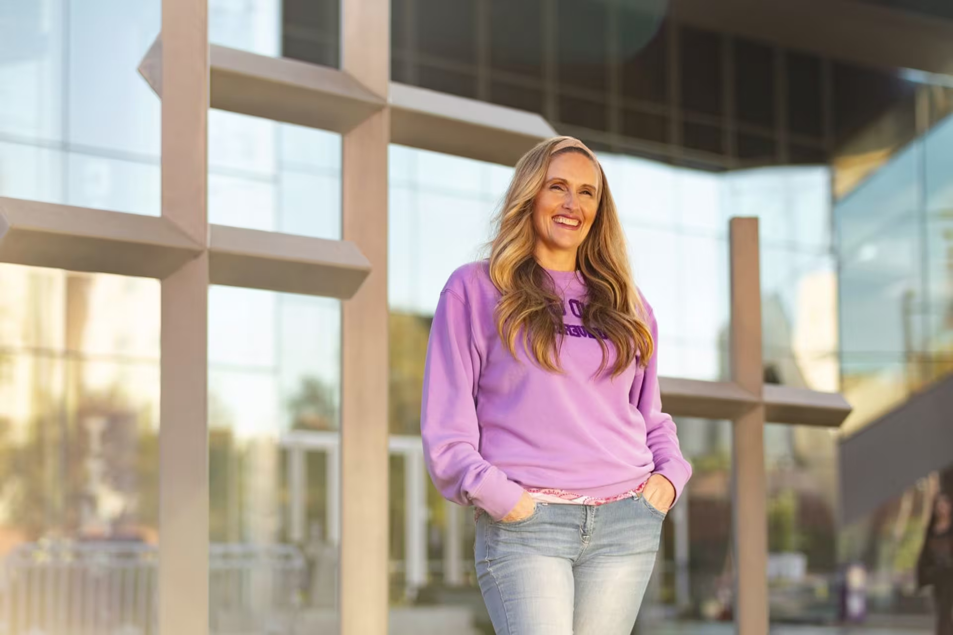 Smiling woman in a purple sweatshirt standing outside a modern building with large crosses, representing faith and community, for the MA in Youth and Family Ministry program.