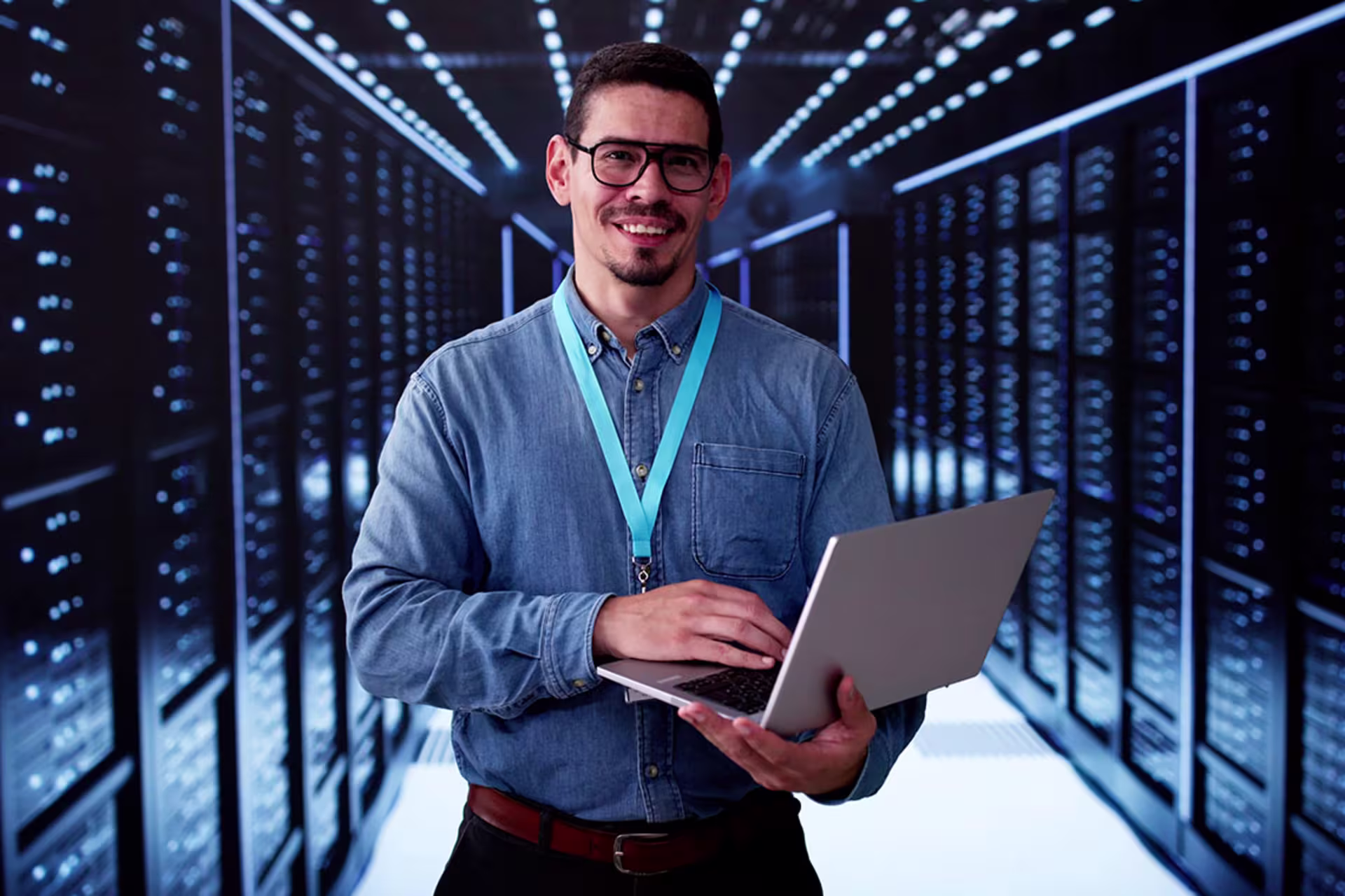 Male engineer in data center server room holding laptop and smiling at camera