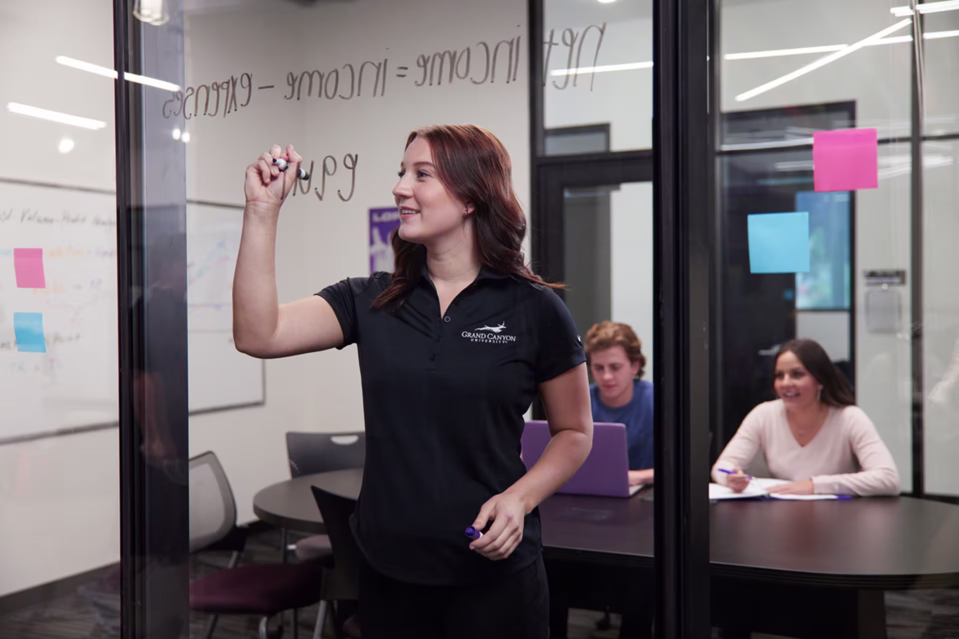 brunette accounting tutor in black gcu shirt writes on glass wall