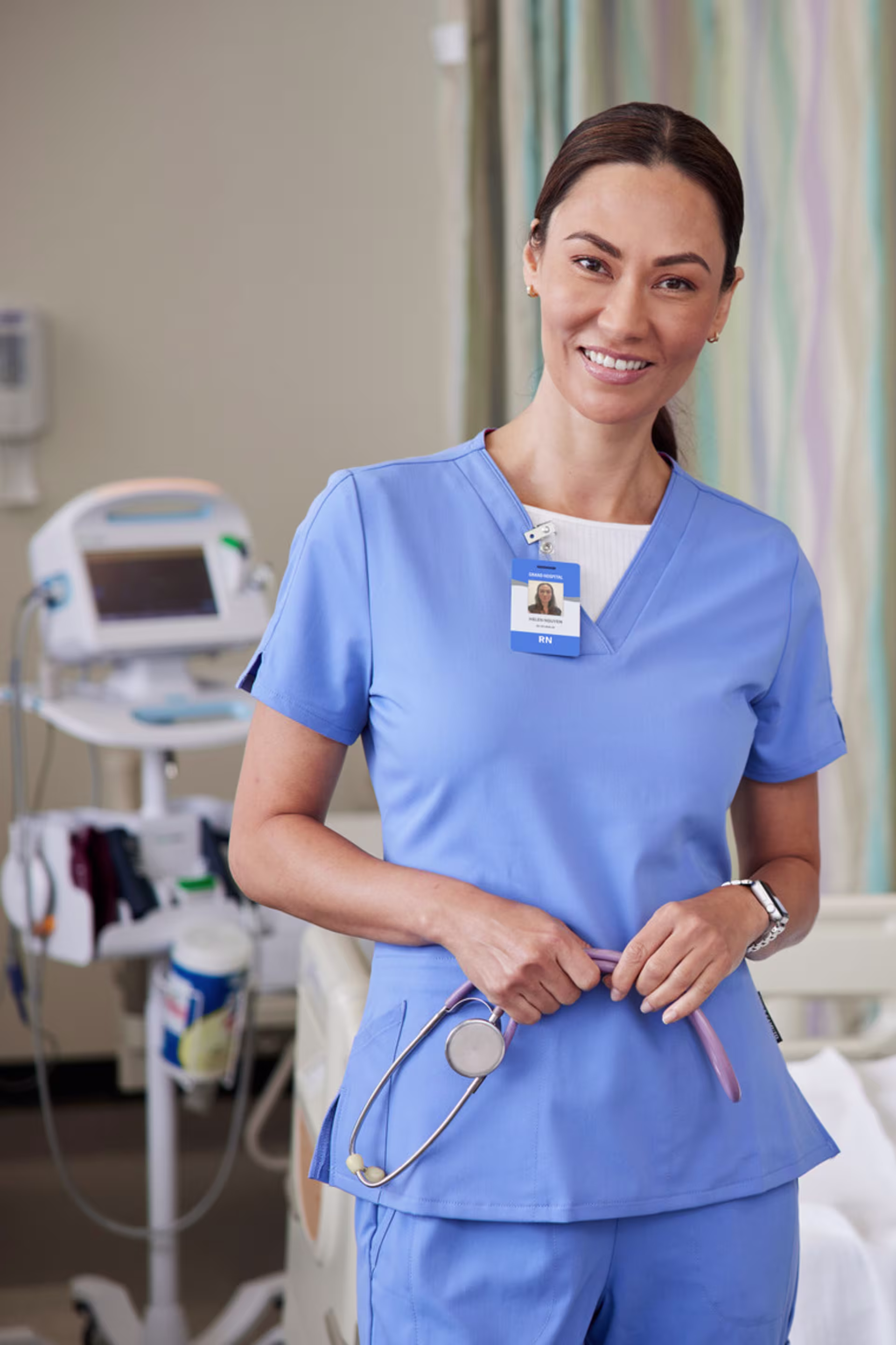 brunette nurse in light blue scrubs holds stethoscope in hospital room