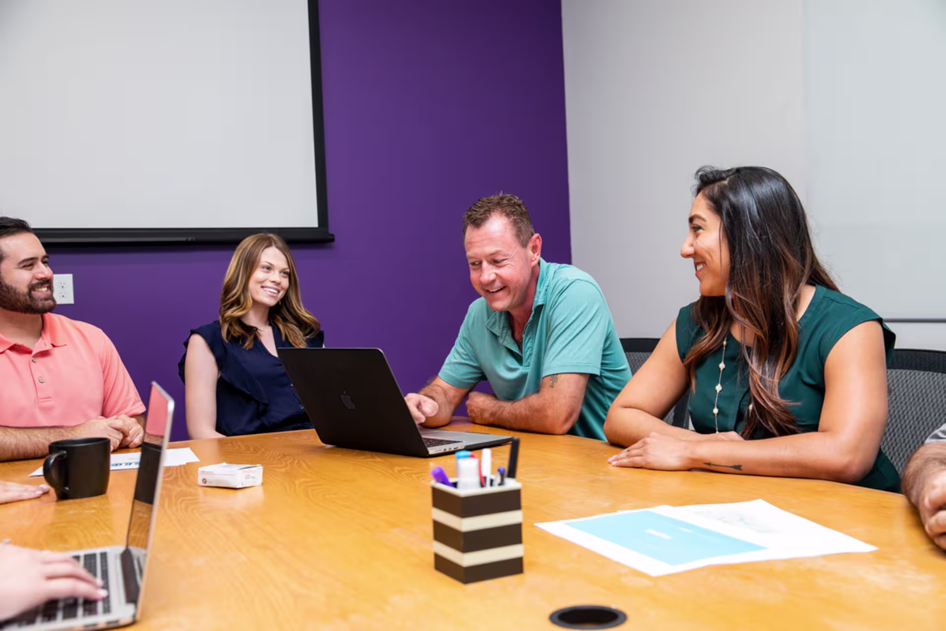 Research group in conference room discussing while man in blue uses laptop.