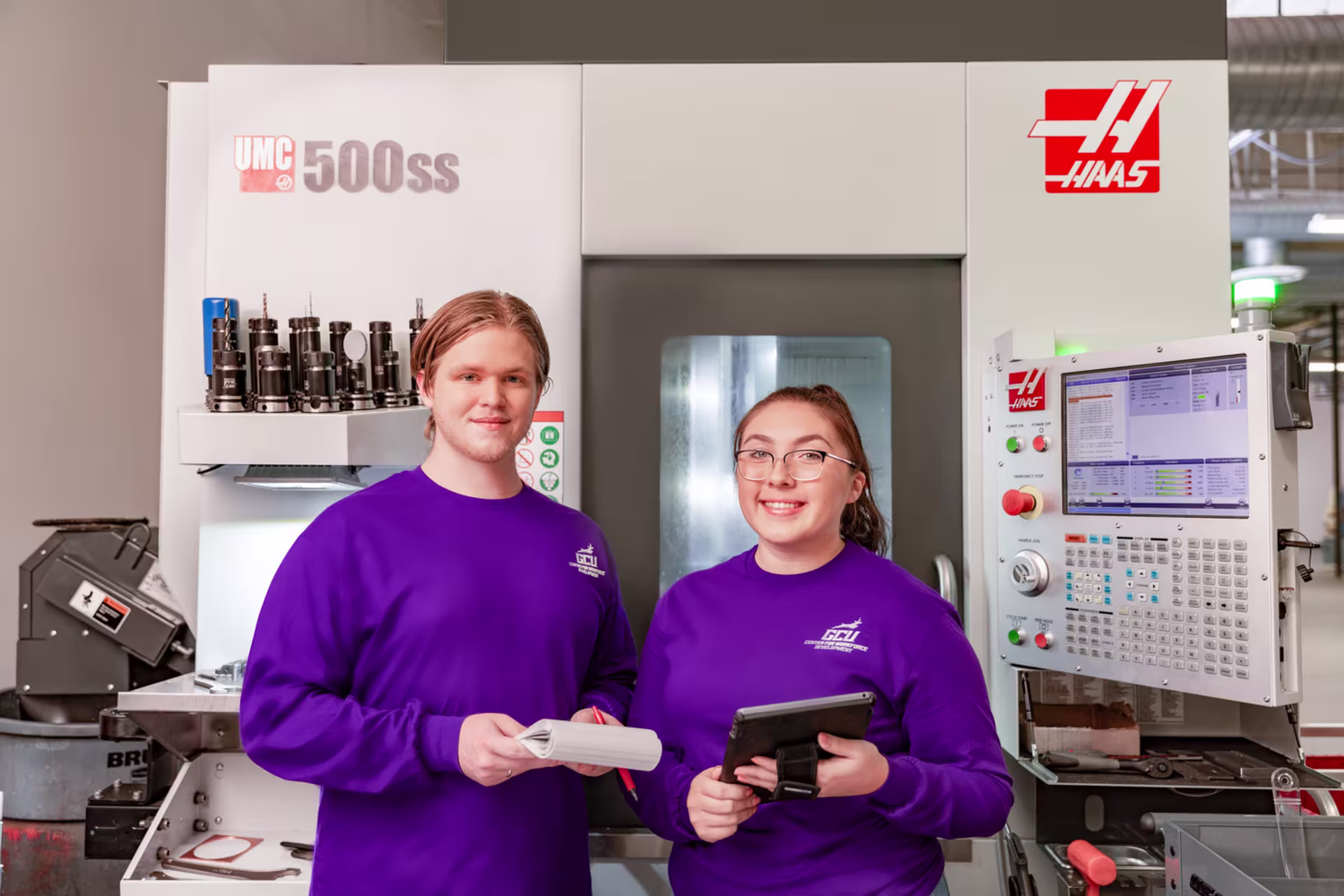 Two GCU students in front of CNC machine smiling while holding a notebook and tablet.