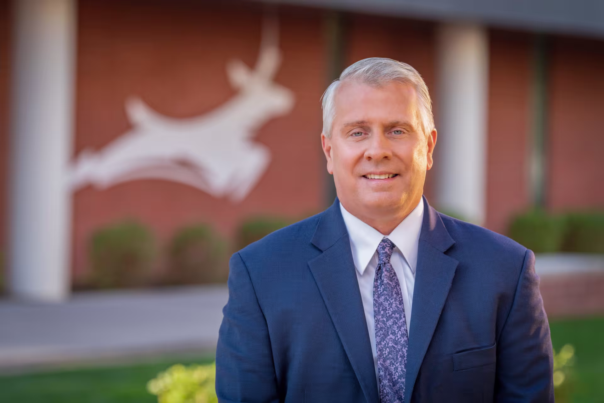 Headshot of James Kossler in front of building with antelope mascot on brick wall.