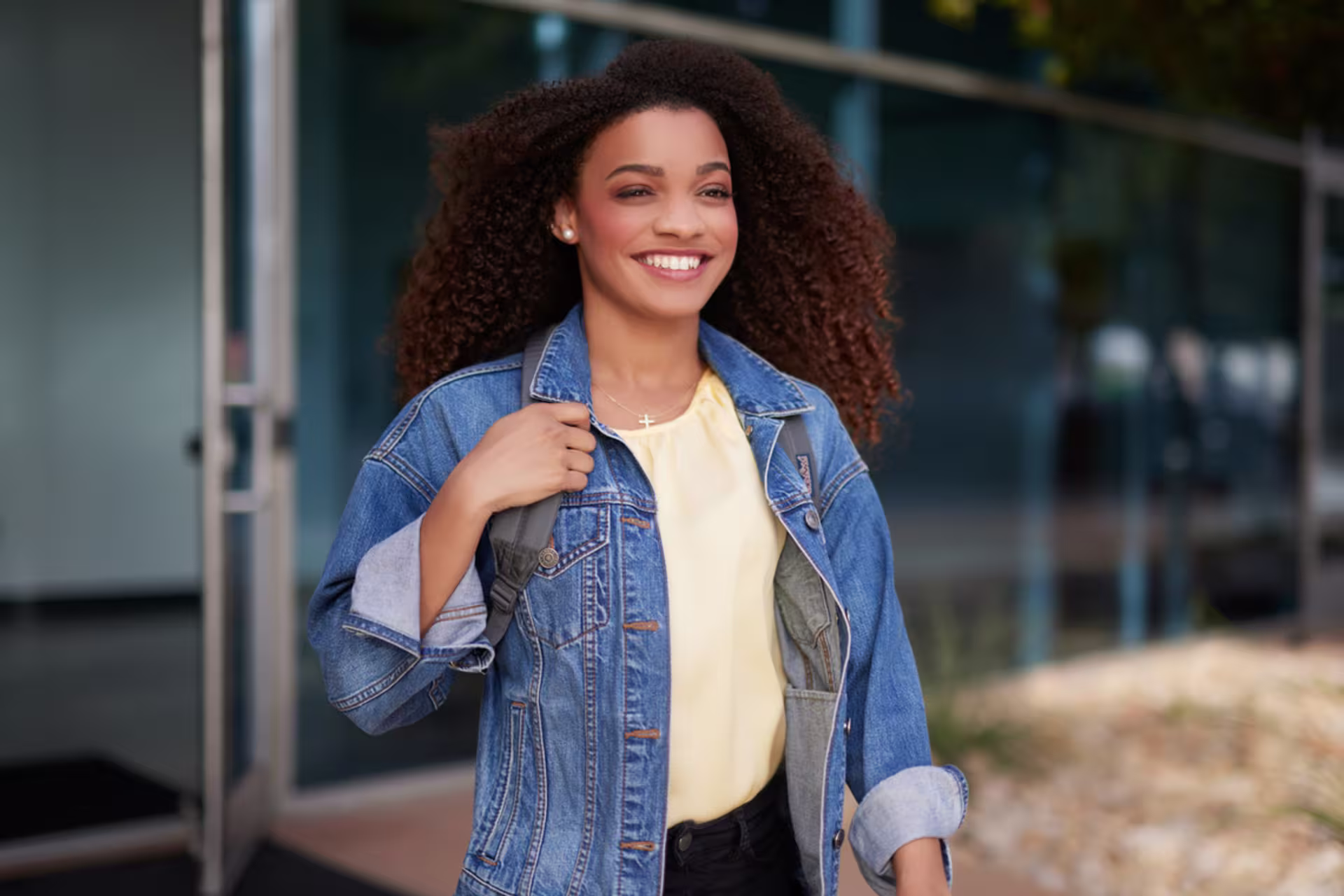 ethnic female student in jean shacket walks out of gcu glass window building smiling