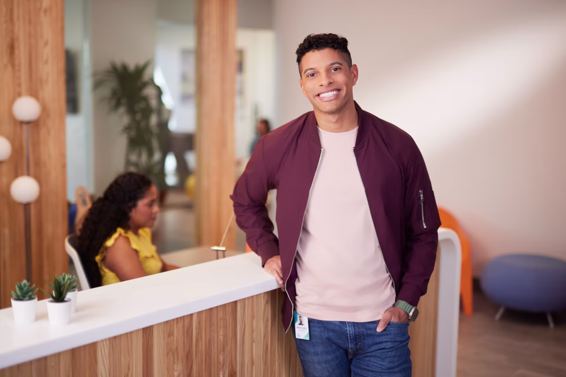 ethnic male student in maroon jacket with jeans stands at assessment office desk with receptionist seated behind him typing